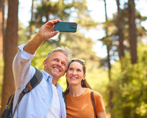 Retired Senior Couple Wearing Backpacks Hiking Through Countryside Posing For Selfie On Mobile Phone - 768624159