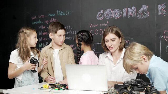 Caucasian teacher using laptop and explain about engineering system while diverse student holding electronic equipment in front of blackboard with prompt or code written in STEM class. Erudition.