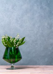 Fotobehang White flowers, lilies of the valley in a green transparent vase, a glass, next to it lies a green scarf and three green balls. Gray background. Plenty of space for text © Alexander