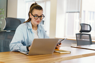 Engaged in multitasking, a poised woman in business casual attire references a digital tablet while...
