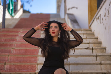 Young, beautiful, brunette woman in an elegant black dress, touching her hair, looking distracted sitting on the stairs of a beautiful white Andalusian village. Concept beauty, fashion, trend, travel