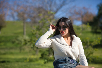 Young, beautiful, brunette woman, wearing a white sweater and a denim miniskirt, sitting on a park...