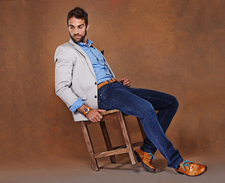 Fashion, serious and man in studio with trendy, stylish and classy suit on chair with confidence. Model, handsome and male person with elegant style for outfit and accessory by brown background.