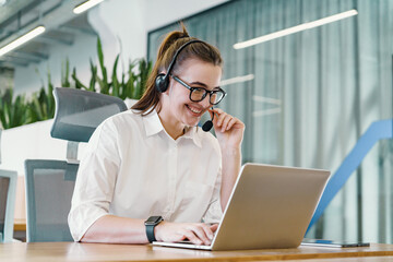 A genial customer support agent with a headset communicates cheerfully on a call while working on a laptop in an office with greenery. - obrazy, fototapety, plakaty