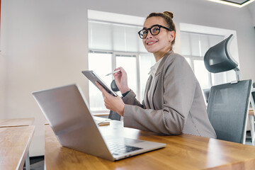 A chic professional woman with a tablet and stylus gives a pleased smile, poised at her desk with a...