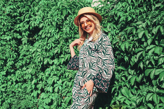 Young beautiful smiling blond hipster woman in trendy summer hippie costume clothes. Carefree female posing in street at sunny day. Positive model outdoors. Cheerful and happy in hat near green bushes
