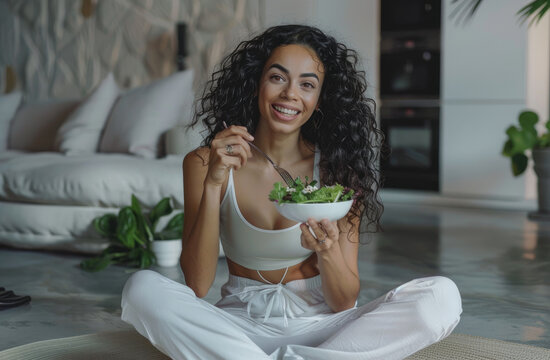 A photo of an attractive woman in her early thirties, wearing white yoga pants and a tank top, sitting on the floor eating salad from a bowl