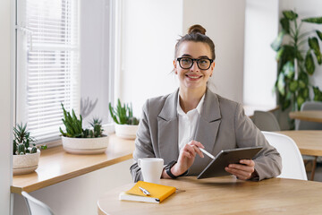 A contented professional with a warm smile handles a tablet in a plant-adorned office, a coffee mug...
