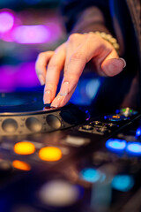 close-up view of a hand adjusting controls on a DJ mixer, illuminated by colorful lights. The fingers, adorned with shiny nail polish, manipulate buttons and knobs.