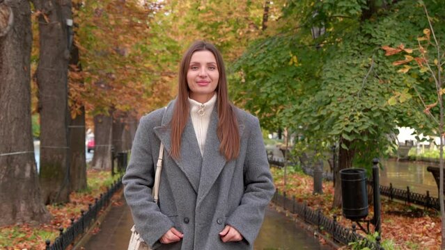 Attractive woman in stylish grey coat walking in autumn park after rain. Camera moving backward