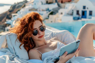 Fototapeten Young red-haired woman in sunglasses using tablet while sunbathing by the pool on Santorini island  © PixelGallery