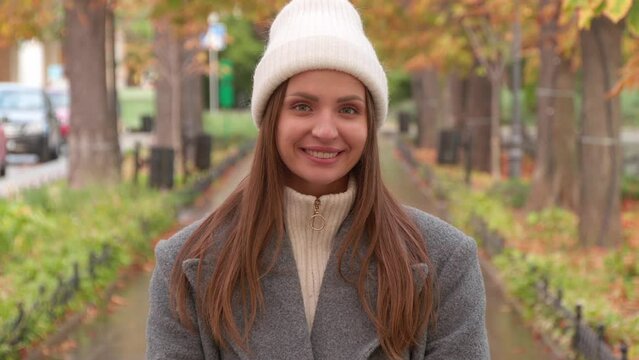 Cute woman in stylish grey coat, warm white hat and mittens in autumn park