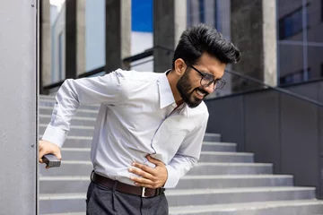 Foto auf Leinwand Suffering from pain, a young Indian male office worker stands outside a building, grimacing and holding his stomach with his hand. © Liubomir