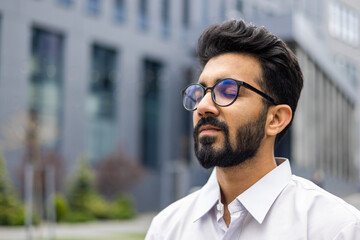 Close-up photo of a relaxed young Indian man wearing glasses and a white shirt sitting outside a...