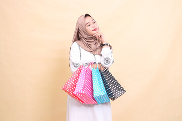 a young Asian Muslim woman wearing a hijab looks left holding her chin and carrying a paperbag shopping bag. for advertising, culture, Eid and Ramadan