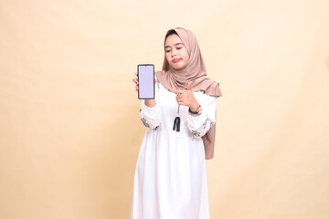 An Asian Muslim girl wearing a hijab frowns candidly, showing a cellphone (gadget) screen pointing to create sales content. for advertising, technology, Eid and Ramadan