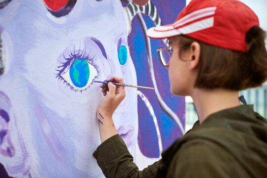 Female painter in red cap draws surreal picture with paintbrush on canvas for outdoor street exhibition, close up side view of female artist apply brushstrokes to canvas, symphony of art creativity
