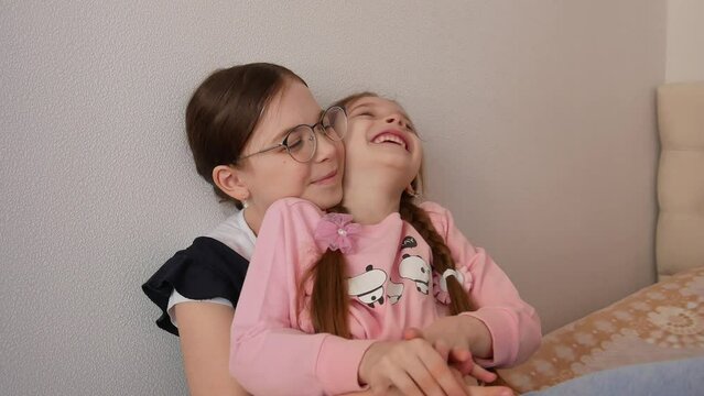 An older sister, a schoolgirl with glasses, holds a younger sister of preschool age in her arms, children smile and tickle, friendly and warm family relations. International Sibling Day.