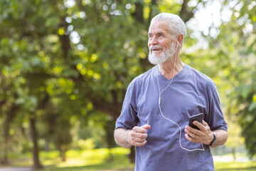 Naklejka na ściany i meble Gray-haired senior man doing sports and running in park, holding phone in headphones, listening to music and smiling.