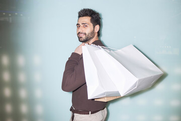 Minimal waist up portrait of smiling bearded man holding shopping bags standing against light blue...