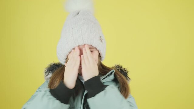 Beautiful little girl in green down jacket and hat. Child covers hands with palms, hides face, plays hide and seek, peekaboo over yellow background.