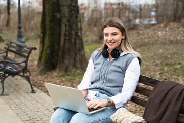 Young successful business woman sitting in park on bench working on laptop computer while listening podcast on wireless headphones. Professional female office worker research outside office building