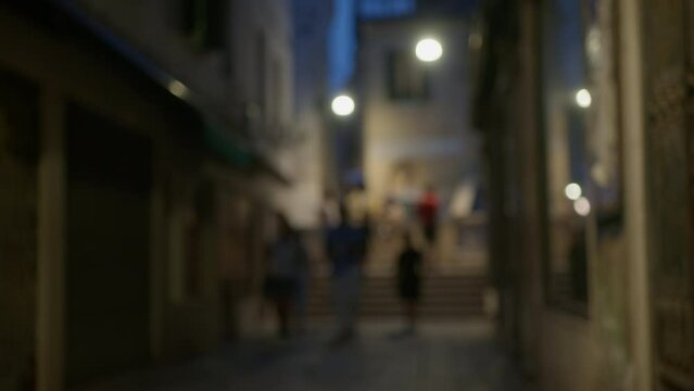 Figures of people walking along a narrow Venetian street, at dusk, illuminated by soft-focus street lights. Blurred for background usage