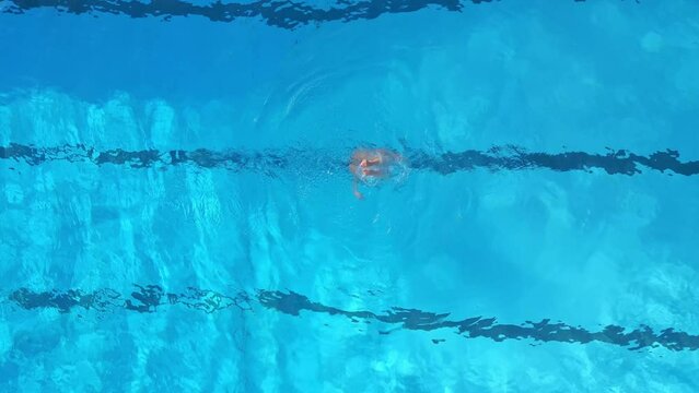A female athlete performs synchronized swimming routines in an outdoor pool, as seen from the air