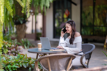 A young woman talking on the phone while working on a laptop in a quiet outdoor cafe.