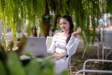 A happy young woman enjoys remote work on her laptop, comfortably seated outdoor of cafe with a coffee.