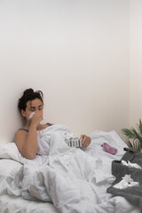 In bed, an ill young woman wipes her nose with tissues, holding medical drops to ease her illness. 