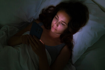 Late at night, a woman rests in bed, drawn to her phone's glow,an image emblematic of insomnia and...