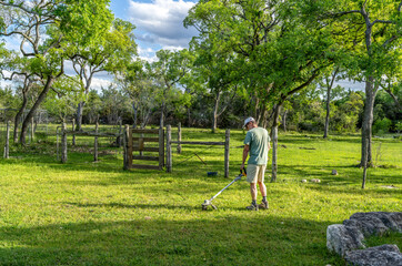 Fototapeta premium Matured Caucasian man using an electric battery operated grass line trimmer in an area with trees, a fence and a gate