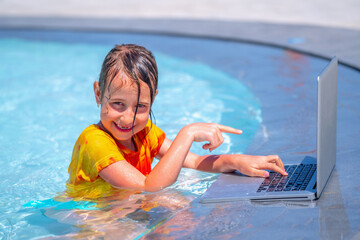 Funny portrait of business child working distance on laptop. Pretty young girl typing on computer in the waterpool. Freelance, remote work on vacation concept.