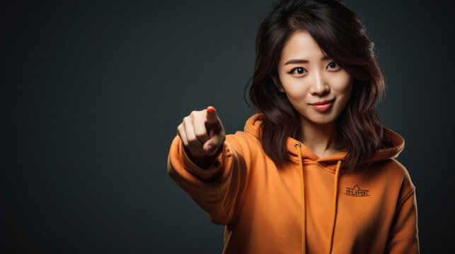 Smiling asian woman in hoodie pointing at camera on black background