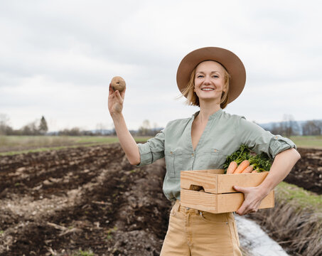 Female farmer holding wooden box full of fresh raw vegetables in agricultural field area. Woman standing proud with freshly bunch harvest outdoors.