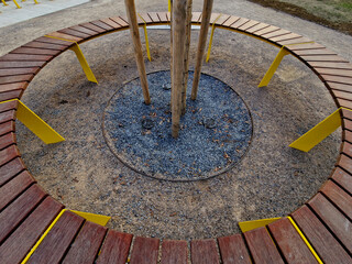 round bench made of wood encloses a tree, sitting around with wood paneling. metal frame. segments made up of planks around the tree. ventilation and watering grids at the roots