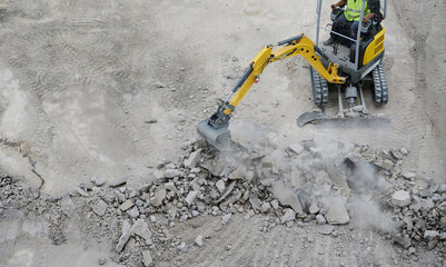 Worker operates a mini excavator to remove old concrete at the ground renovation work at the construction site. - obrazy, fototapety, plakaty