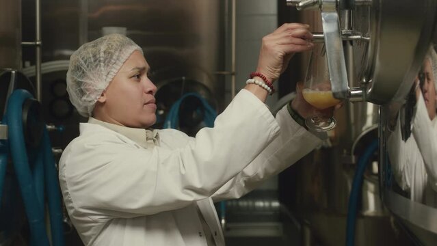 Waist up of Biracial female brewery technician in white lab coat taking sample of freshly brewed beer from fermenter tank, checking its appearance and taste