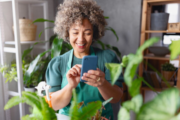 Mature mixed race smiling woman gardener working in home garden, using plant identification app for mobile phone