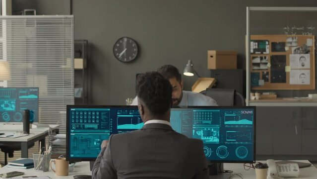 Medium shot of ethnically diverse male cybersecurity officers coworking in agency, exchanging folders with confidential data info and monitoring network system on multiple computer displays