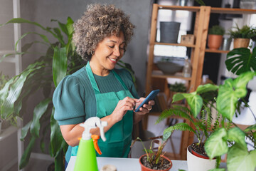 Mature mixed race smiling woman gardener working in home garden, using plant identification app for mobile phone
