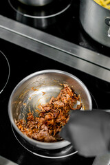 Close-up of a person in black gloves cooking food in a stainless steel pan on a modern stove in a well-equipped kitchen