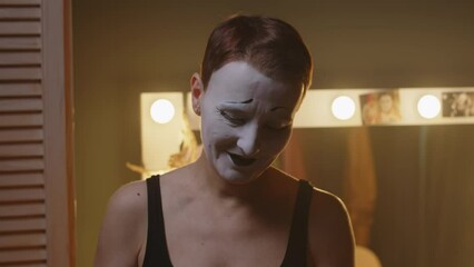 Zoom-in shot of happy adult mime actress wearing stage makeup looking through fans letters after concert, leaning on mirror table in backstage room with yellow dimmed light