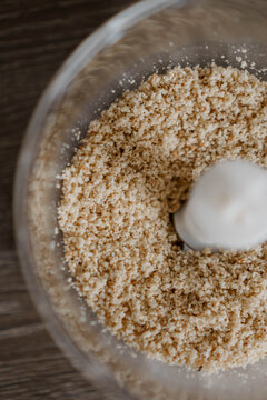 This photograph features finely ground hazelnuts in a food processor with a white spinning blade, set against a wooden tabletop. The ground nuts have a coarse texture.