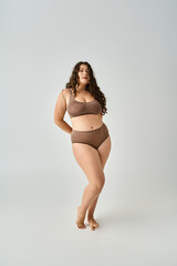 charming curvy woman in underwear posing with hands behind back in grey background