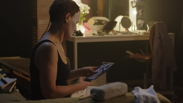 Medium shot of mature mime actress looking at photos of herself performing on stage at different times while sitting on couch in backstage room after concert