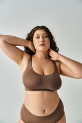 Obraz premium seductive curvy girl in brown underwear with curly hair touching hands to neck and looking to camera
