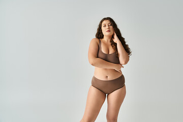 alluring plus size young woman in brown lingerie with curly hair posing with arms