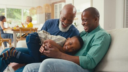 Grandfather with father and grandson playing and laughing on sofa at home with rest of family in background - shot in slow motion - Powered by Adobe